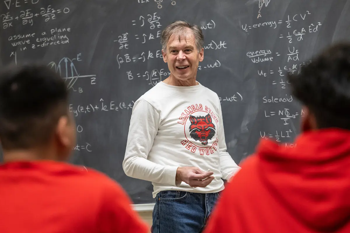 Professor teaches physics in front of chalkboard filled with equations, engaging with students in class.