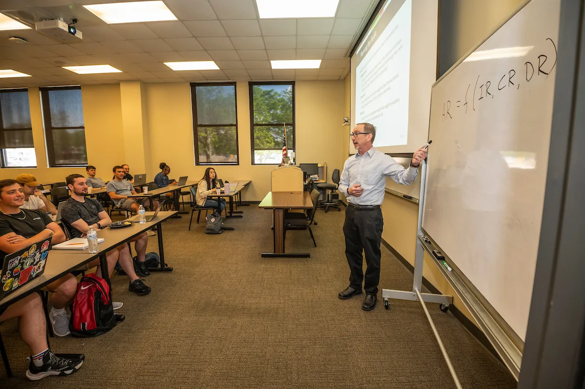 Professor teaches class using whiteboard and screen while students listen and take notes.