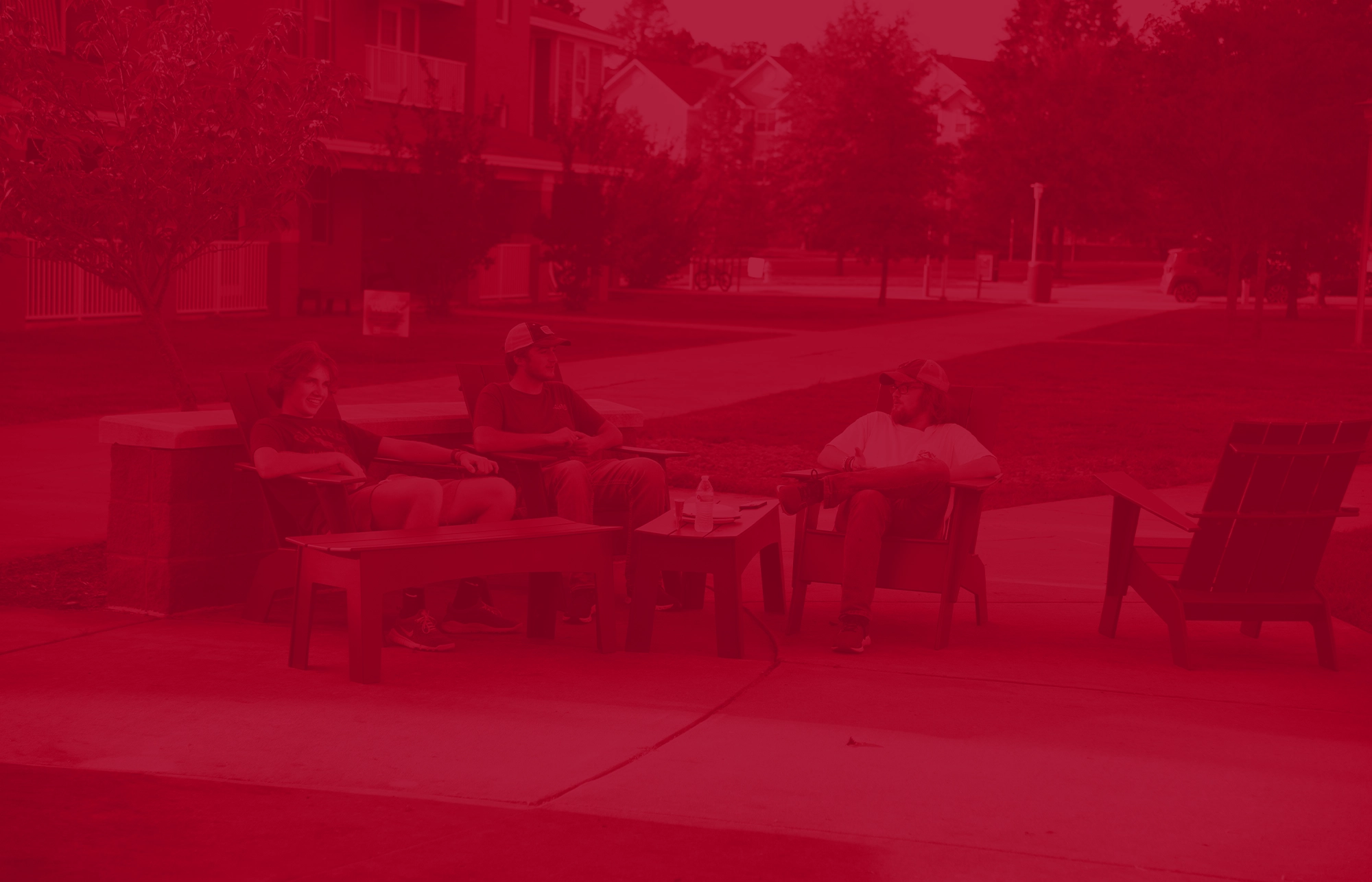 Students seated in outdoor lounge chairs around a low table on a paved patio area, with apartment buildings and landscaped greenery in the background, shown with a red color overlay.