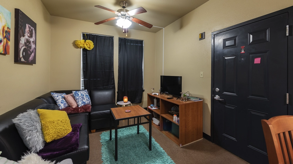 View of Red Wolf Den living room from kitchen area, showing black sectional sofa, coffee table, teal rug, TV stand, and front door with ceiling fan overhead.