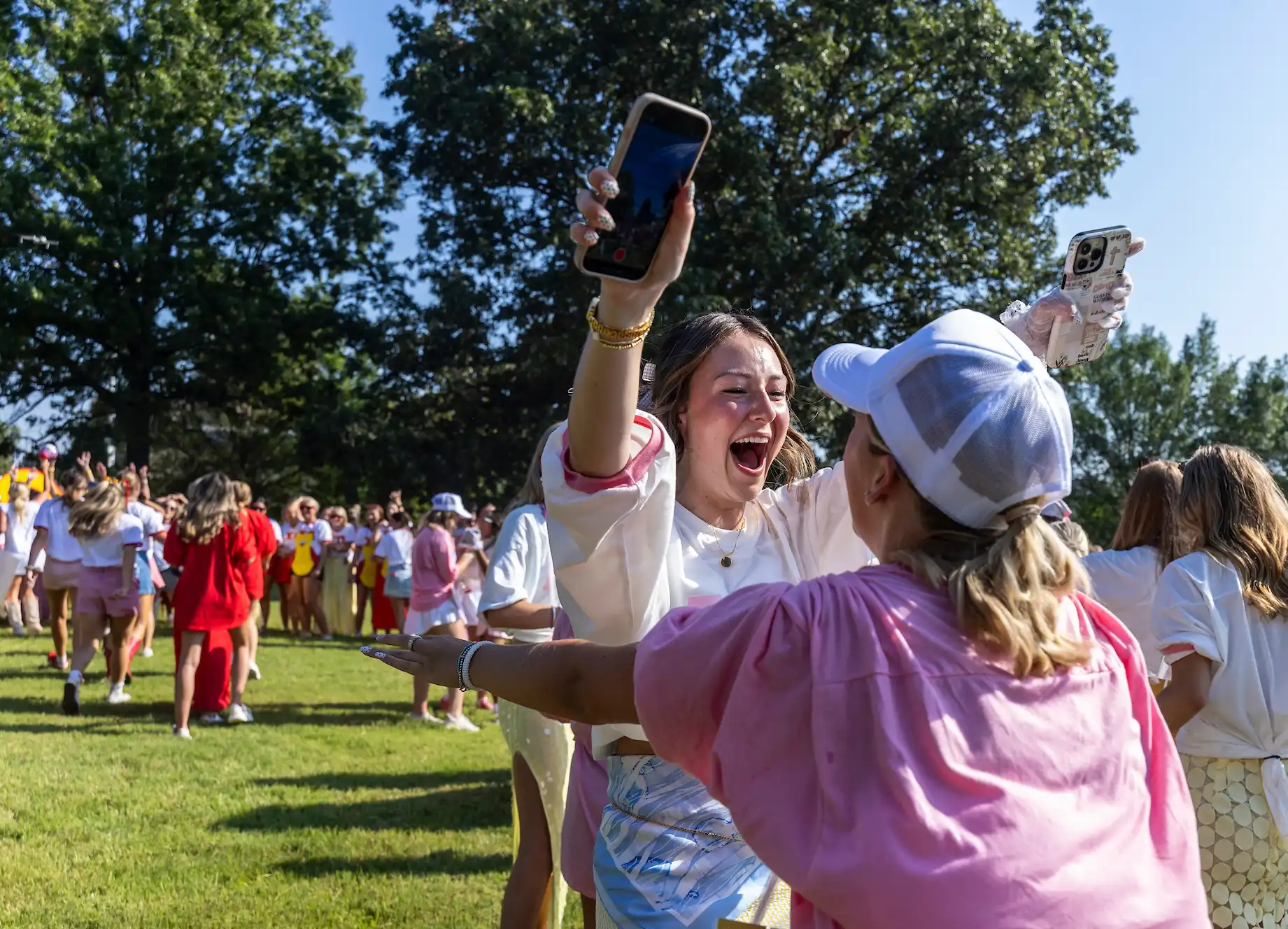 Girl holding cell phone leaning in for hug with another girl outside on grassy lawn at Sorority Bid Day.