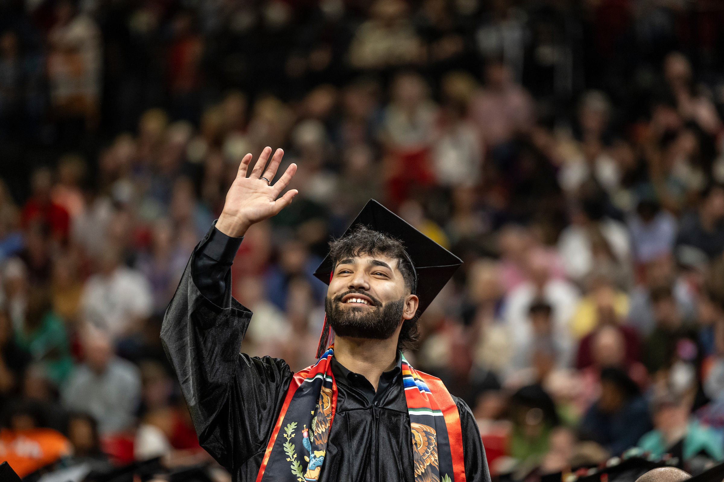 Smiling graduate in cap and gown waves during commencement ceremony.