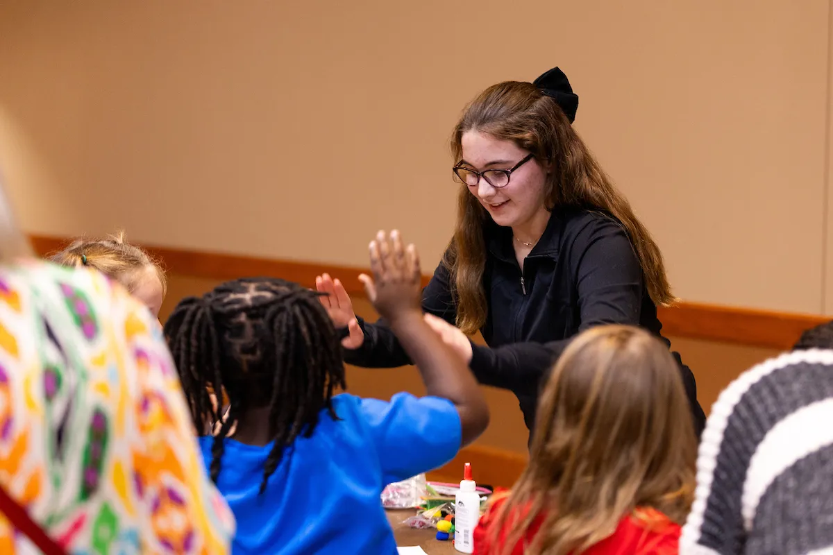 A student gives a high five to a group of elementary students who are gathered around a table.
