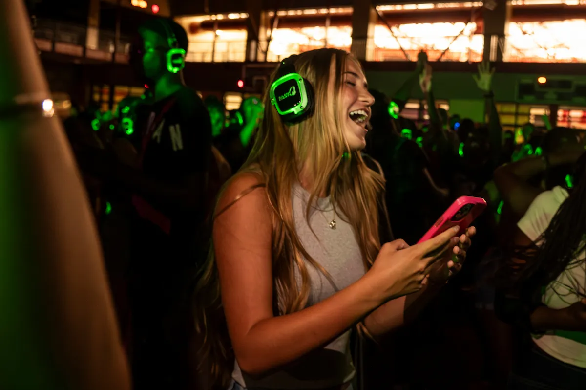 Student wearing glowing headphones laughs and dances at a silent disco with friends on campus.