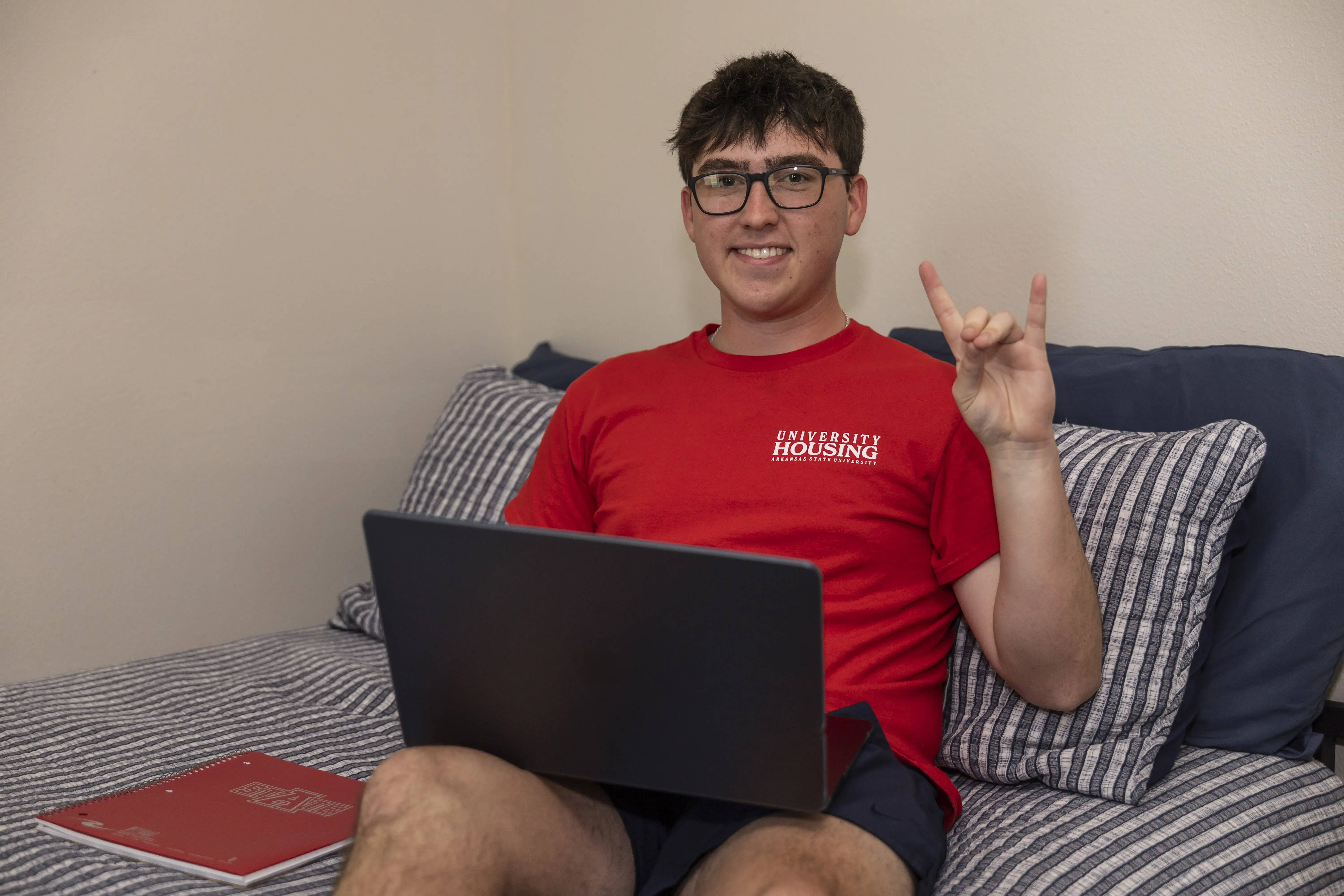 Student sitting on bed with laptop and making a 'wolves up' motion with hand.