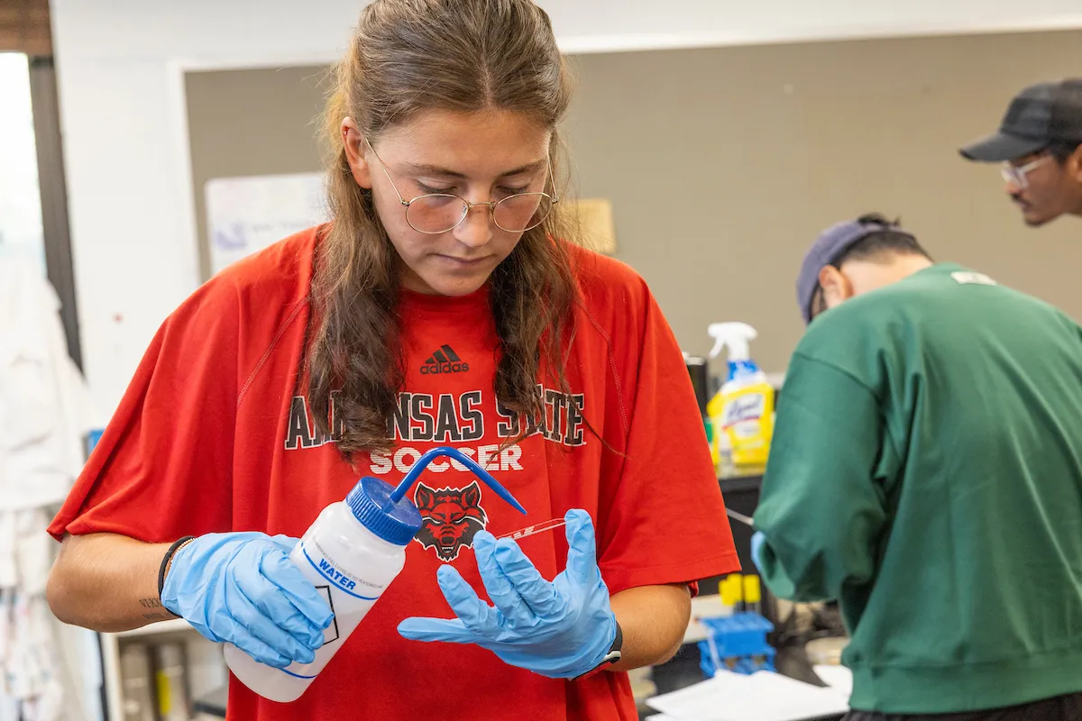 A student in gloves and a Red Wolves shirt works with a squeeze bottle in a science lab.