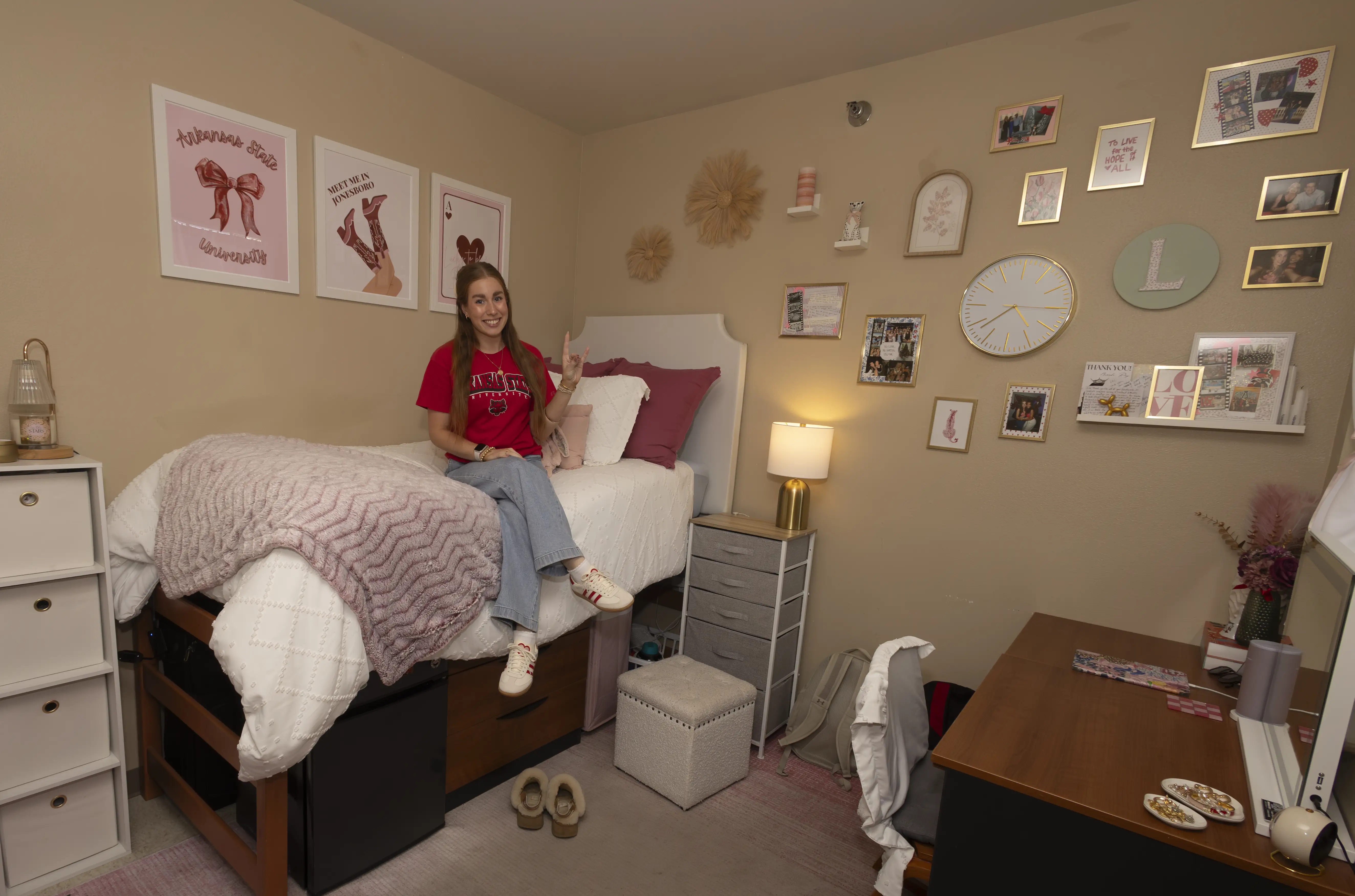 Student sitting on bed in decorated residence hall room, holding their hand up in a 'wolves up' motion.