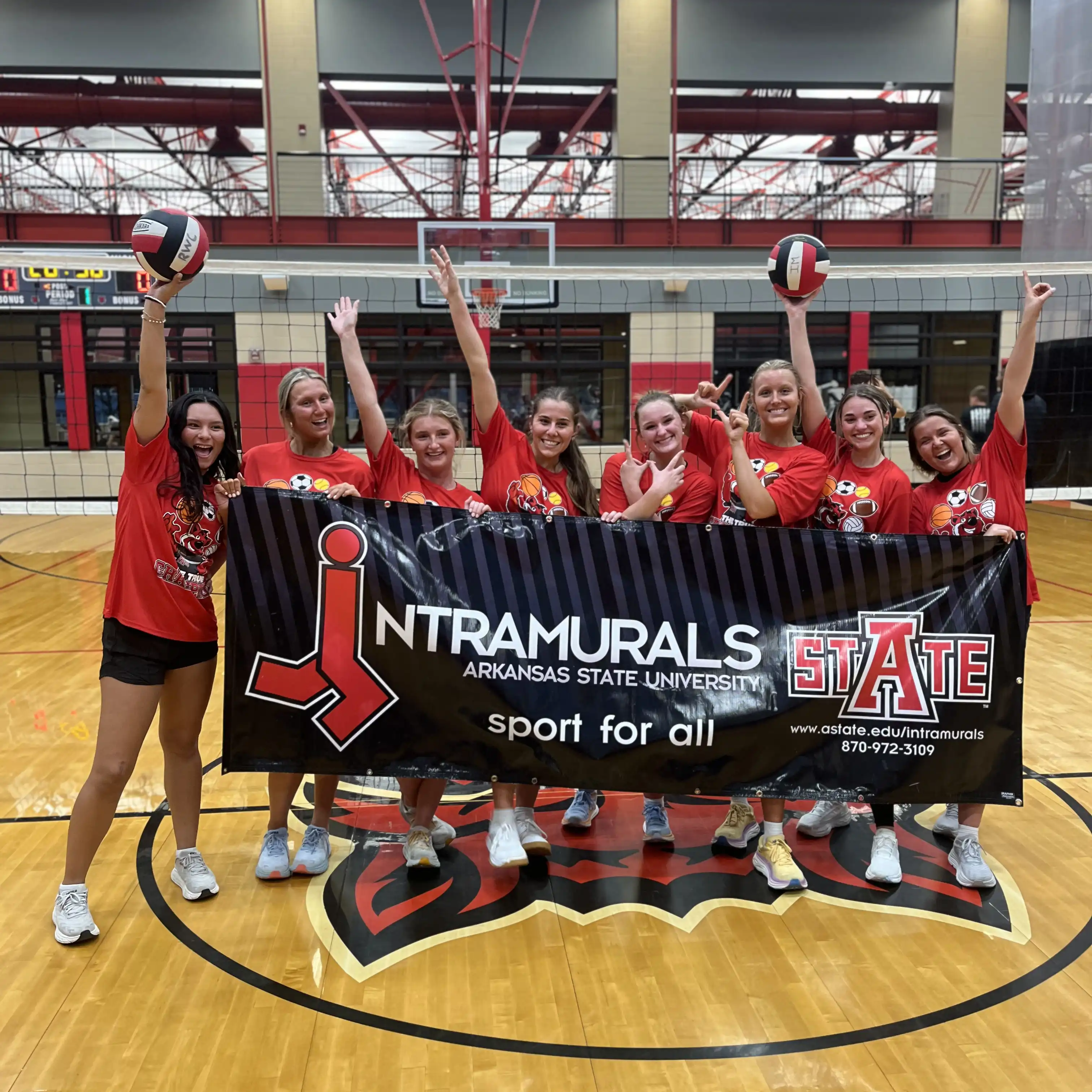 Students celebrate with championship banner and volleyballs in gym.