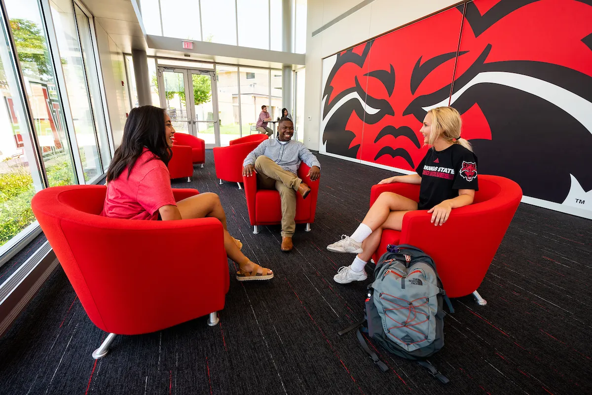 Three students chat in red chairs near a large Red Wolves mural inside a bright campus lounge.