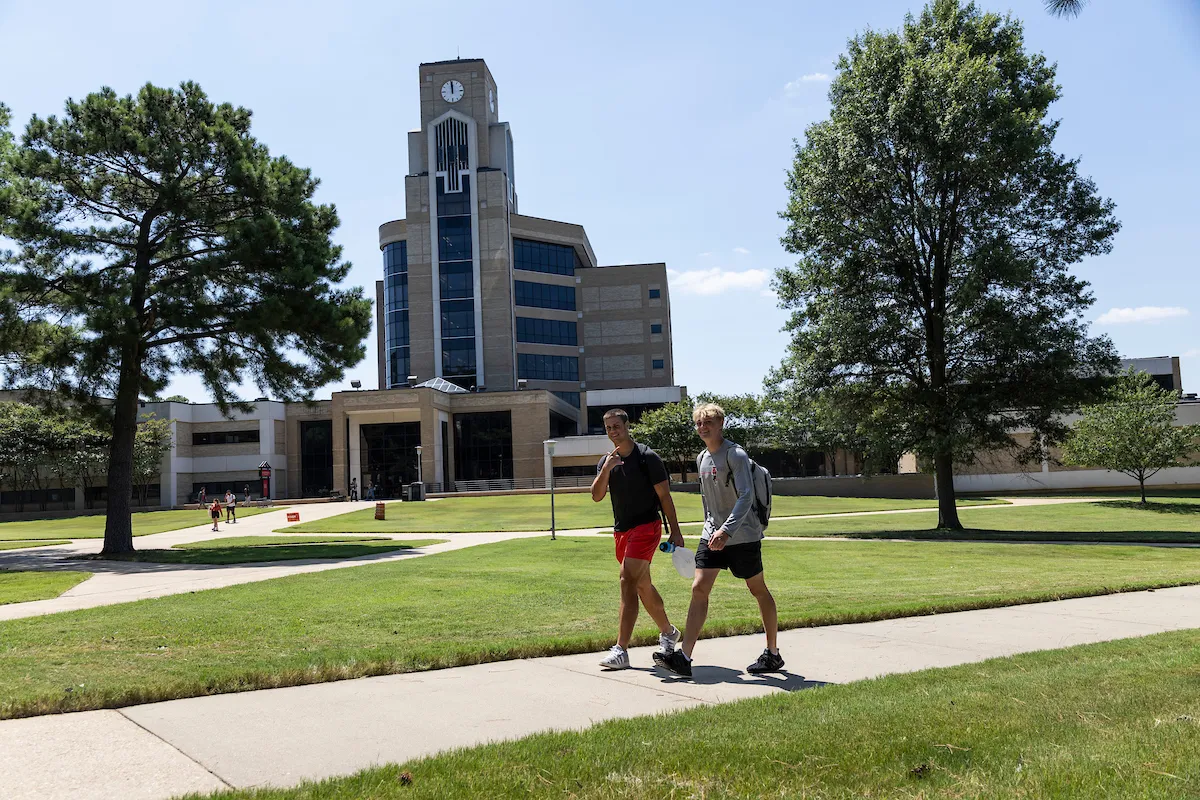 Two male students walk across a sunny A State campus sidewalk in front of the Dean B. Ellis Library clock tower.