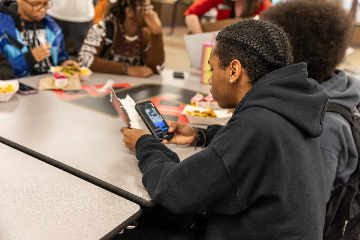 Student at a table looks at phone while eating, surrounded by others socializing in a casual dining setting.