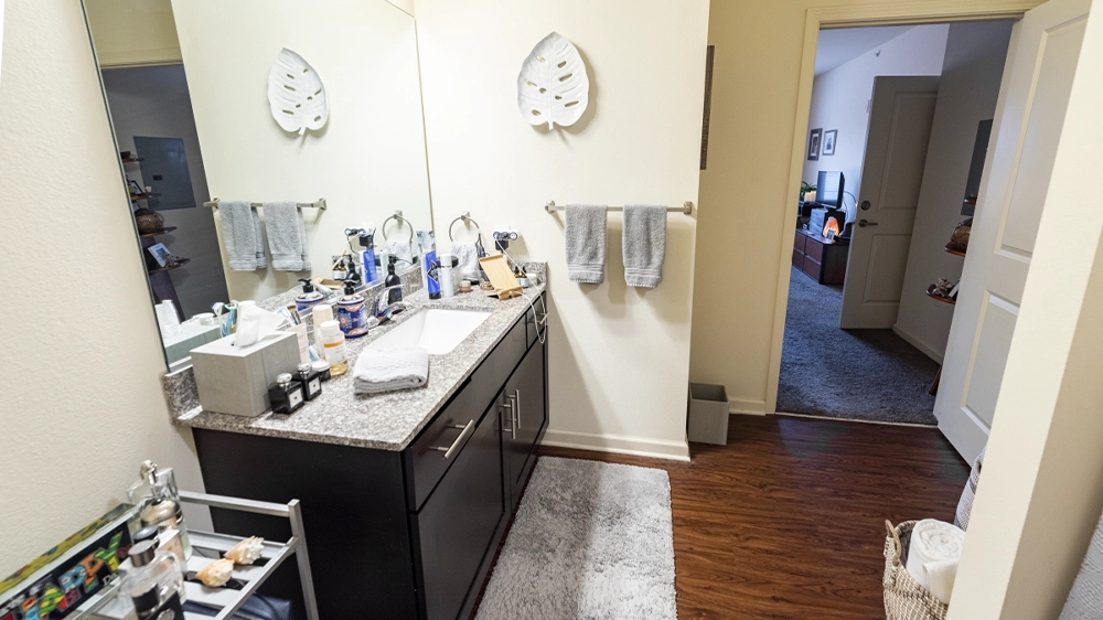 Bathroom with granite countertop vanity, large mirror, and dark wood cabinets, leading to a hallway with view of the bedroom.