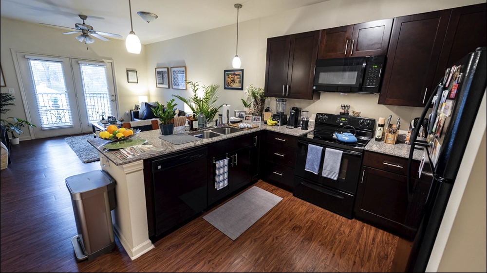 Open kitchen with granite counters and dark wood cabinets overlooking a living area with hardwood floors and glass doors to a balcony.