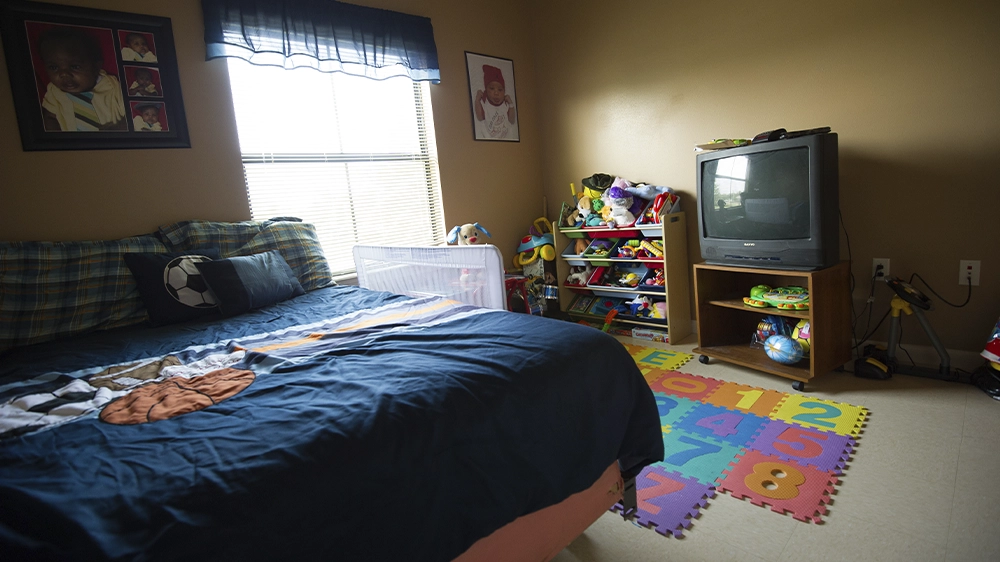 Bedroom with a twin bed featuring sports-themed bedding, a colorful foam play mat, toy shelves, and a TV stand near a window with blinds.