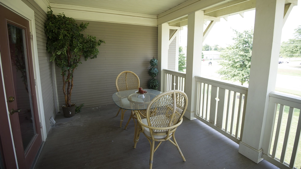 Covered porch with wicker chairs and a glass-top table, surrounded by railing and greenery.