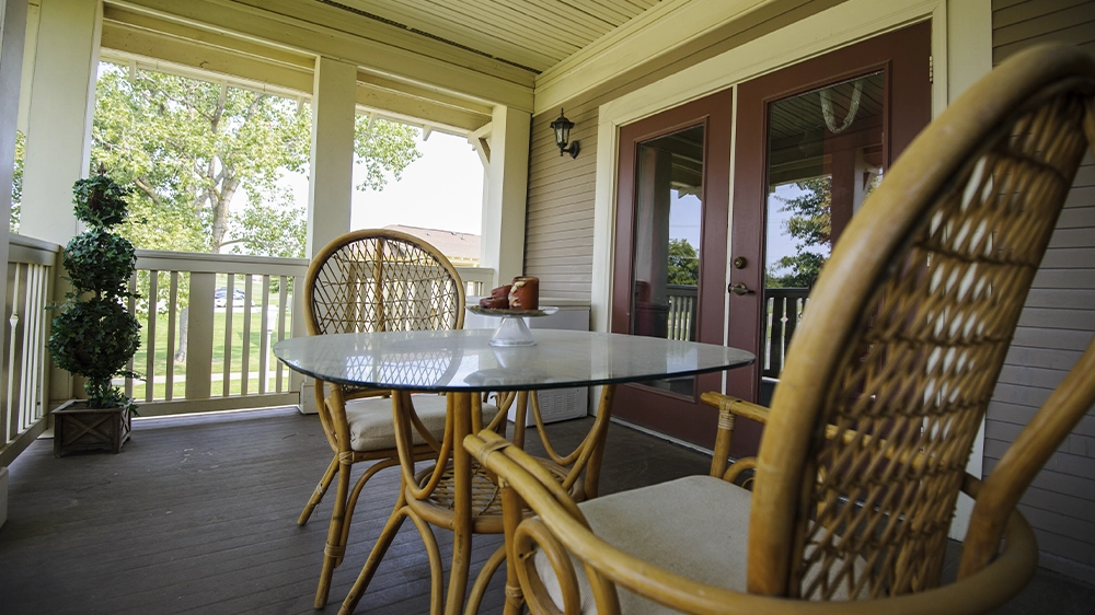 Covered porch with a glass-top table and two wicker chairs, overlooking green lawns and trees.