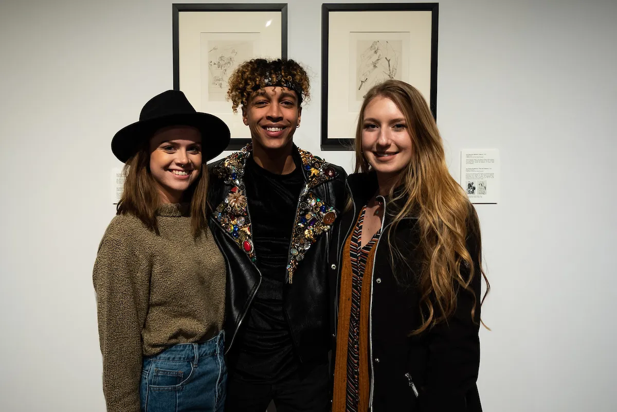 Three students pose in front of framed artwork during a campus art gallery event.