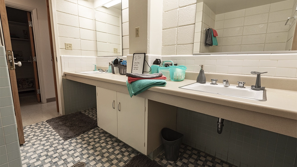 Suite-style bathroom with two sinks, large mirrors, and a beige countertop. Cleaning supplies and toiletries are placed on the counter, and the floor features a patterned tile design.
