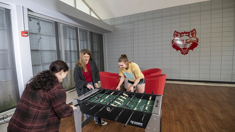 Three people playing foosball in a bright lounge area with wood-style flooring, large windows, and a gray wall featuring a red wolf logo.