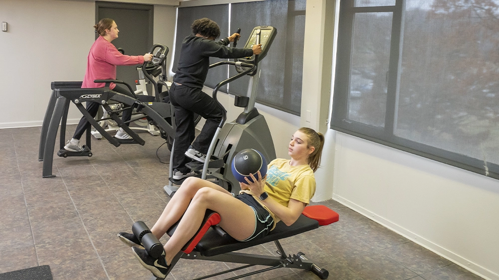 Fitness room with two cardio machines and a workout bench near large windows, offering space for exercise and wellness.