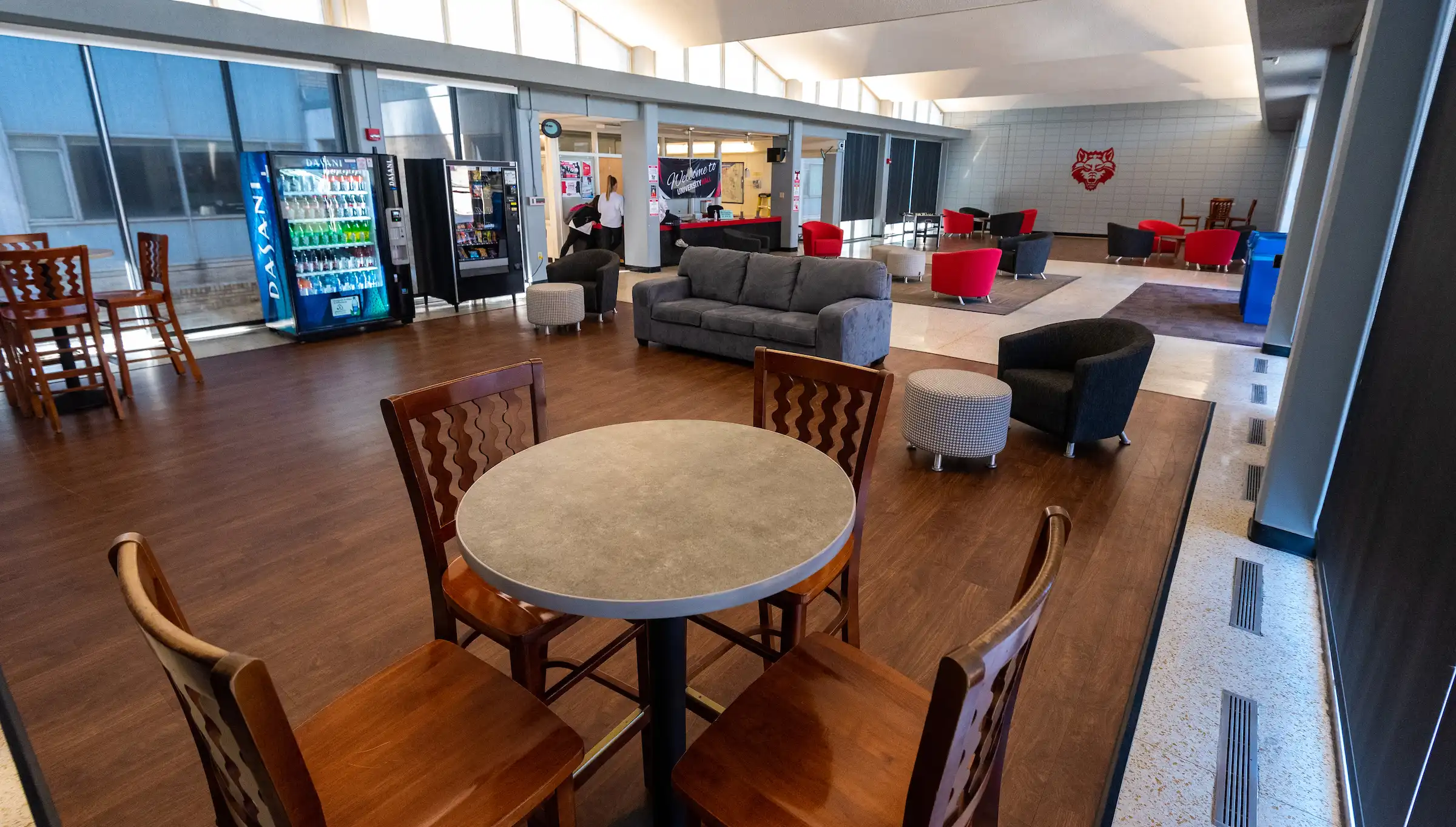 Spacious lobby inside University Hall featuring a gray sofa, round ottomans, vending machines, and large windows with black shades. Several people are visible near the back of the room, and the space includes bright overhead lighting and wood-style flooring.