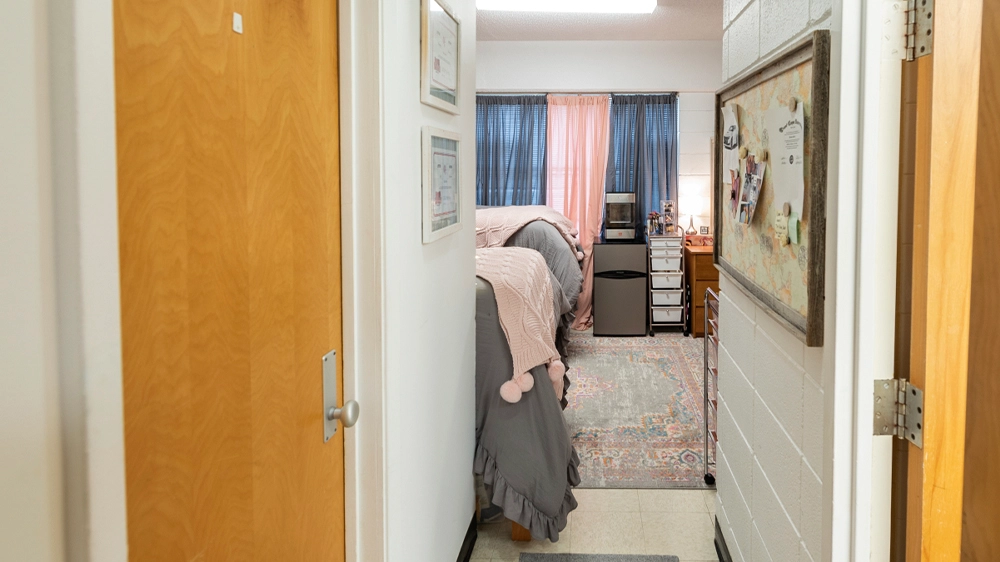 View from the entry hallway into a University Hall bedroom featuring twin beds with gray and pink bedding, a colorful rug, and a window with curtains.