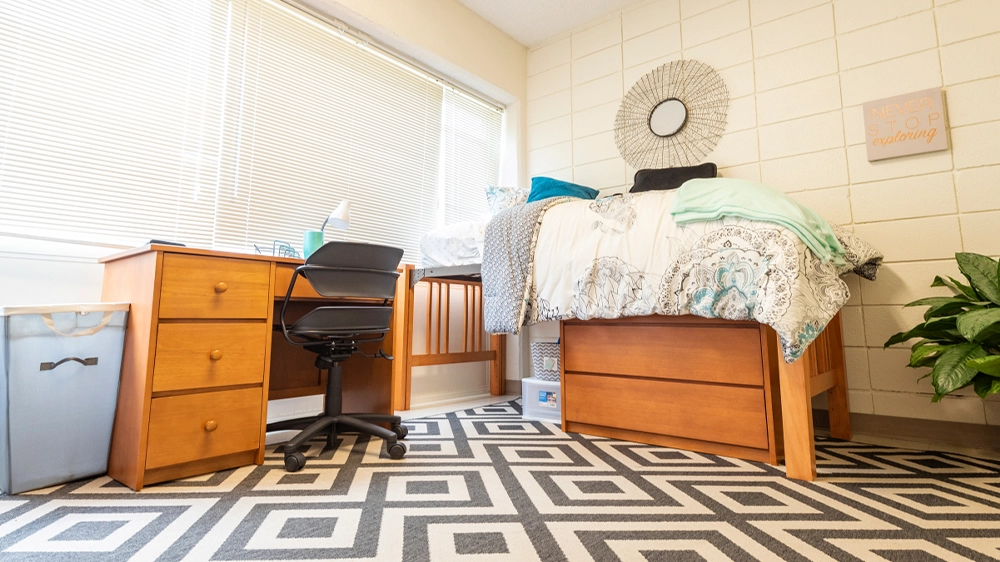 Low-angle view of a lofted bed with patterned bedding, a wooden desk and chair, and a decorative wall mirror above the bed.