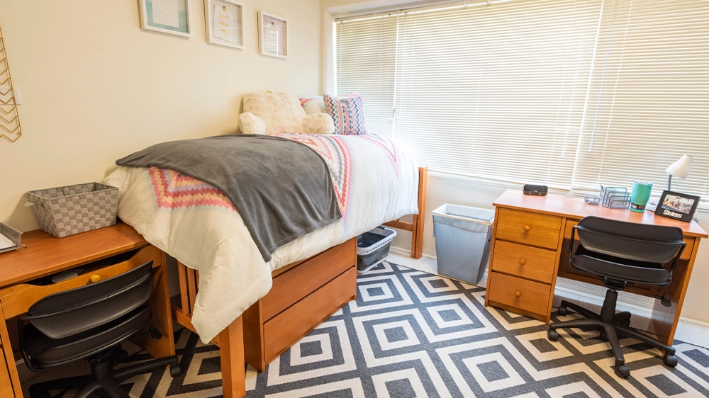 Bedroom layout featuring two lofted beds with patterned bedding, two desks with chairs, and a large window with blinds.