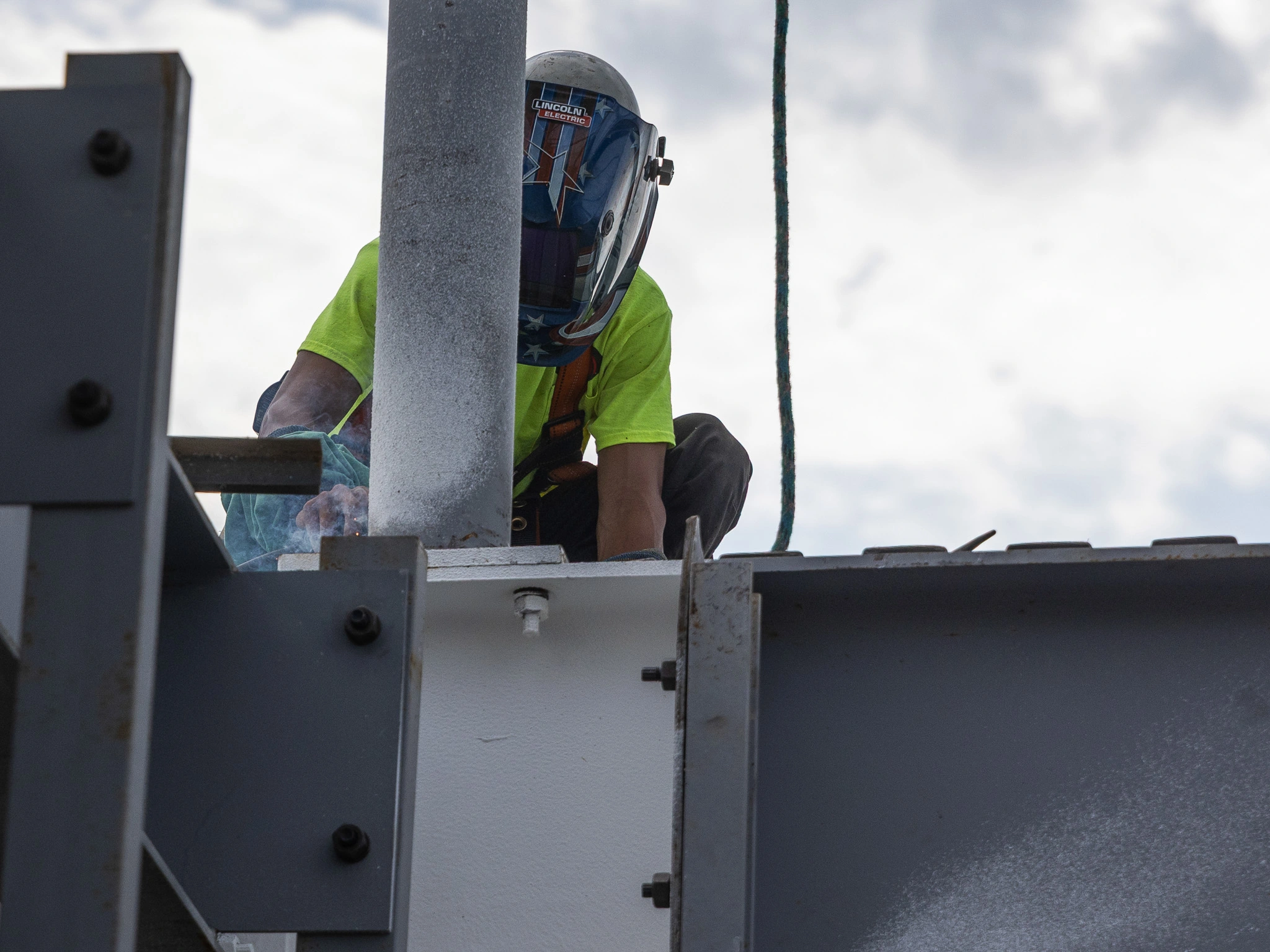 “Person wearing a welding helmet and safety gear works on a steel structure outdoors, standing on a platform next to metal beams and equipment.”