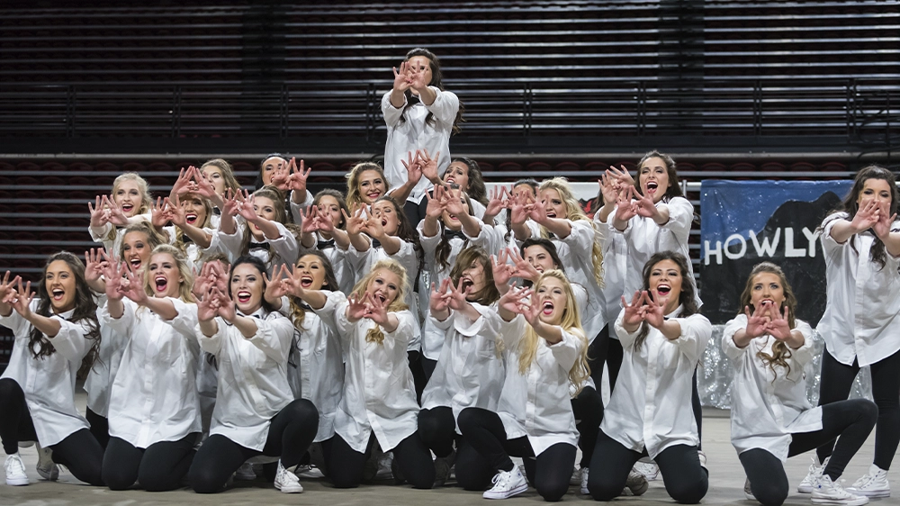 Group of performers in white button-down shirts and black pants posing in a choreographed formation during a step show on an indoor stage.